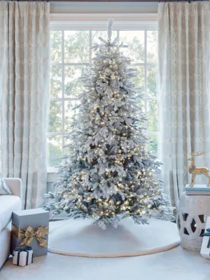 Pre-lit Queen Flock LED Lux Full Artificial Christmas Tree glowing with warm lights in front of a window.
