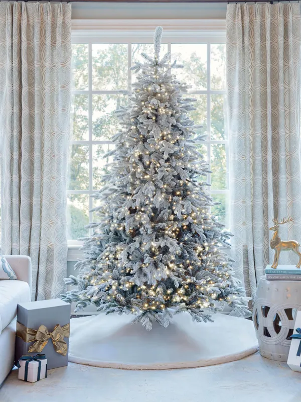 Pre-lit Queen Flock LED Lux Full Artificial Christmas Tree glowing with warm lights in front of a window.