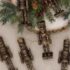BronzeNutcracker Christmas Jungle six bronze nutcracker glass ornaments arranged on linen near pine branches and berries.
