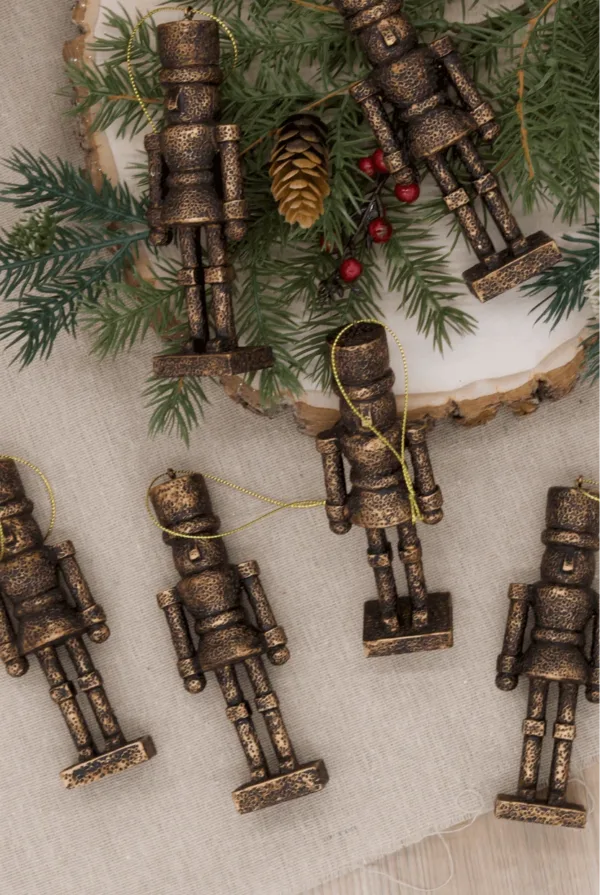BronzeNutcracker Christmas Jungle six bronze nutcracker glass ornaments arranged on linen near pine branches and berries.