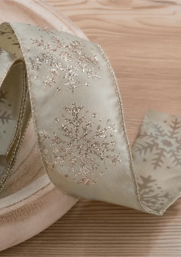 Close-up of champagne-colored Forest Snowflake Ribbon with glittery snowflake patterns coiled on a wood surface.