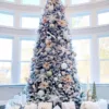 Tall King Flock Full Artificial Christmas Tree decorated with blush pink poinsettias and white ornaments in front of arched windows.