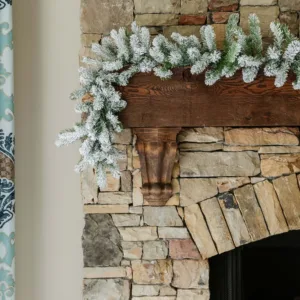 King Flock Garland with snow-flocked evergreen branches draped across a rustic dark wood mantel on a stone fireplace.