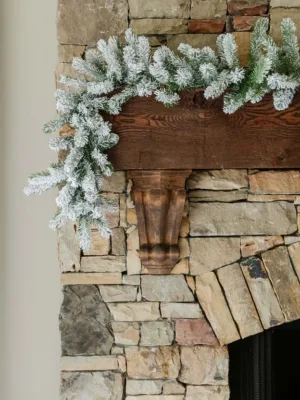 King Flock Garland with snow-flocked evergreen branches draped across a rustic dark wood mantel on a stone fireplace.