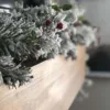 Close-up of frosted King Flock Garland branches accented with bright red berries along a light wood mantel.