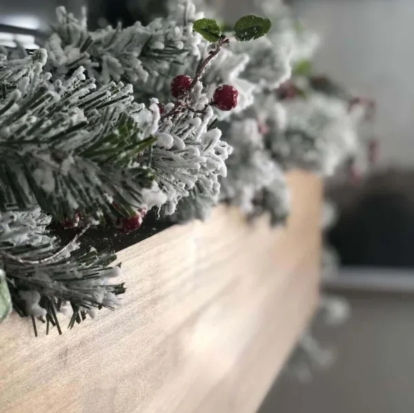 Close-up of frosted King Flock Garland branches accented with bright red berries along a light wood mantel.