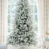 Pre-lit King Flock Full Artificial Christmas Tree with warm white lights standing on a white tree skirt in front of a window.
