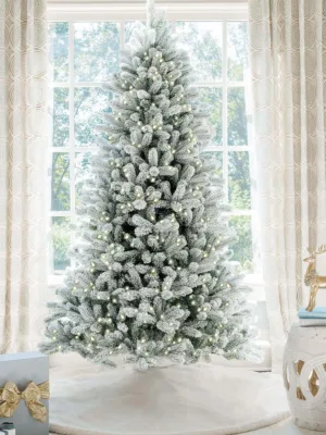 Pre-lit King Flock Full Artificial Christmas Tree with warm white lights standing on a white tree skirt in front of a window.