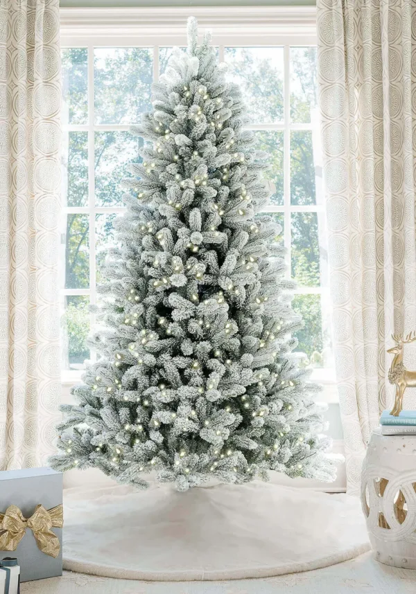 Pre-lit King Flock Full Artificial Christmas Tree with warm white lights standing on a white tree skirt in front of a window.