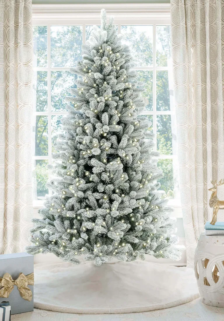 Pre-lit King Flock Full Artificial Christmas Tree with warm white lights standing on a white tree skirt in front of a window.