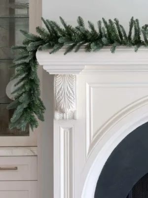 King Noble Fir Garland with blue-green fir branches draped over the edge of a white fireplace mantel.