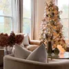 Pre-lit Queen Flock Full Artificial Christmas Tree decorated with bronze ornaments and wide champagne ribbon next to a leather armchair.