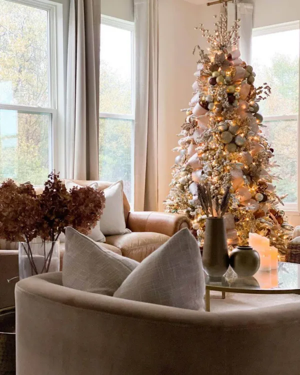 Pre-lit Queen Flock Full Artificial Christmas Tree decorated with bronze ornaments and wide champagne ribbon next to a leather armchair.
