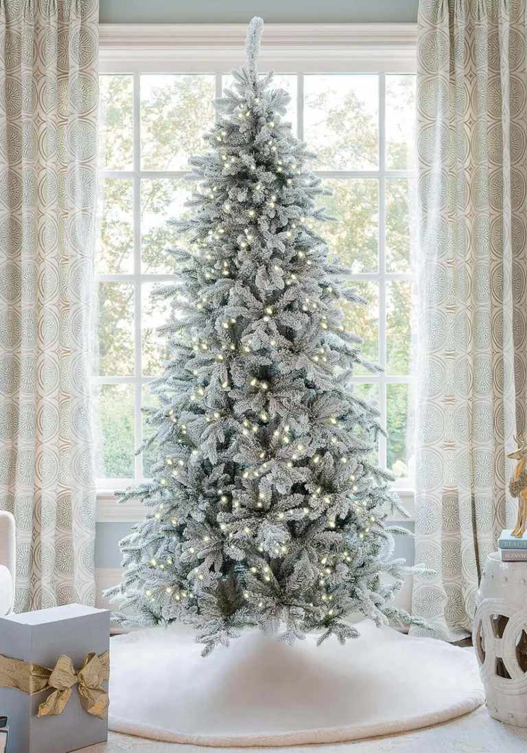 Pre-lit Queen Flock Slim Artificial Christmas Tree glowing in front of a window on a white tree skirt.
