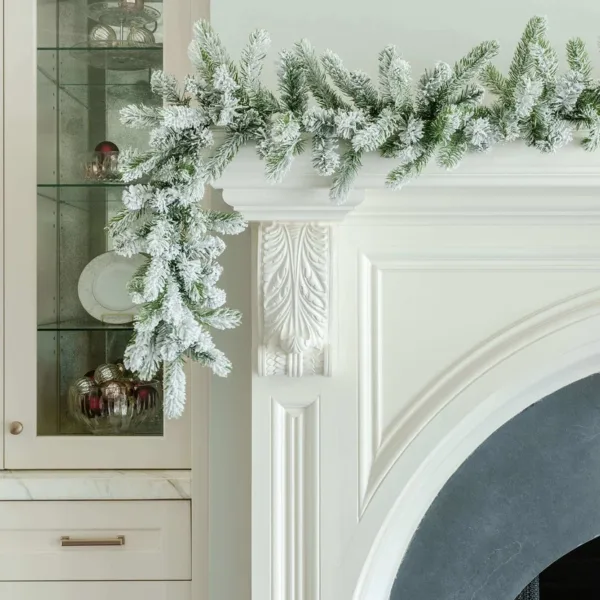 King Flock Garland with snow-dusted pine foliage cascading over the edge of a white fireplace mantel next to glass cabinetry.