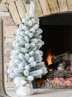 3' King Flock Artificial Christmas Tabletop Tree in a burlap base standing on a stone fireplace hearth next to a lit fire.