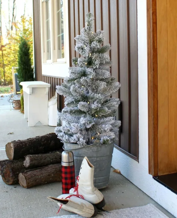 3' King Flock Artificial Christmas Tabletop Tree placed in a metal bucket on a porch next to ice skates and firewood.