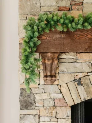 Royal Fir Garland with lush green evergreen branches draped across a rustic dark wood mantel on a stone fireplace.